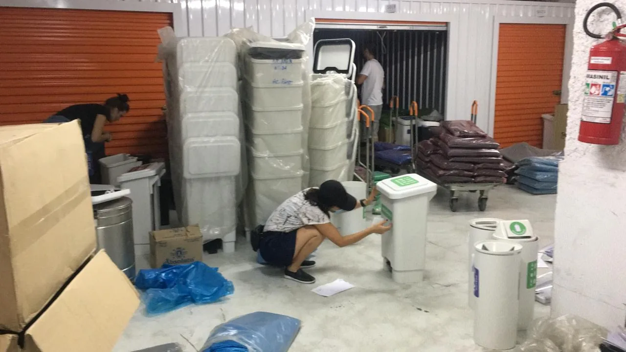 Yuki crouching to apply a label to a waste bin at the warehouse, among stacked bins and bags